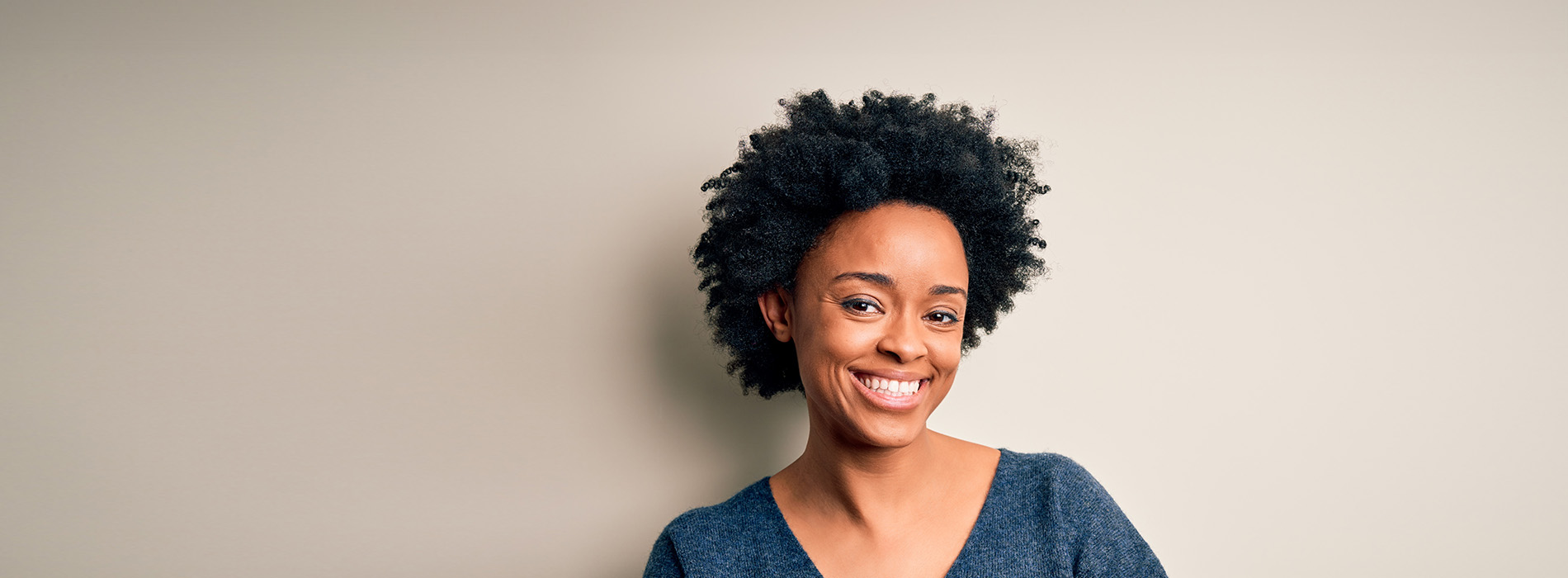 The image is a photograph of a woman with light skin, smiling at the camera. She appears to be in her late twenties or early thirties and has long hair. Her eyes are looking directly at the camera, and she is holding up her index finger near her mouth as if she s making a point or emphasizing something. The background is plain and light-colored, which suggests that this could be a stock photo used for various purposes such as advertising, personal branding, or lifestyle content.