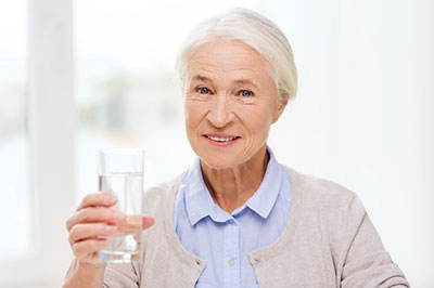 An elderly woman holding a glass of water, smiling and looking directly at the camera.